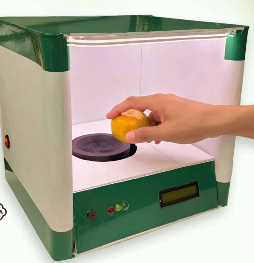 The WaKaBe grading box: a hand placing an orange on the load cell platform inside the lit chamber, with an LCD and three lamps on the front panel